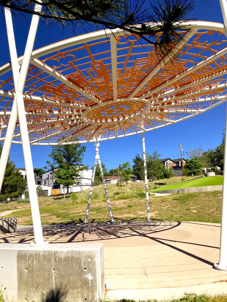 Underside view of a large circular orange metal canopy sculpture with intricate cutout patterns depicting city buildings and clouds, supported by white poles over a park plaza.