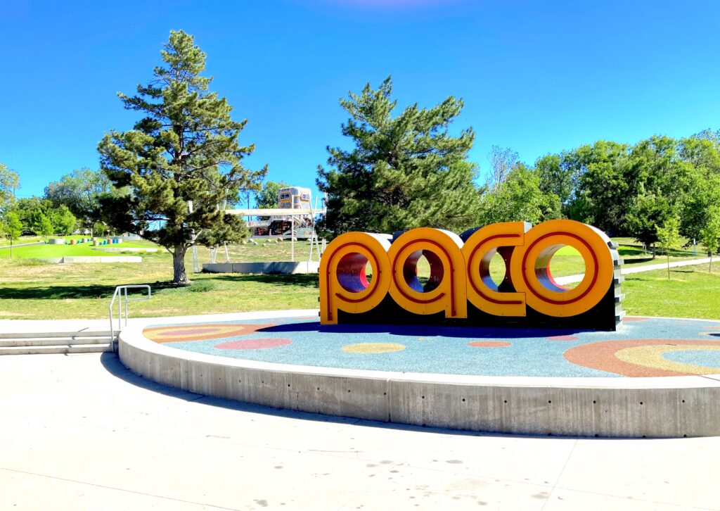 Large yellow letters spelling "PACO" with cutout tunnel holes for climbing, set on a colorful circular play surface with trees and a slide structure in the background.