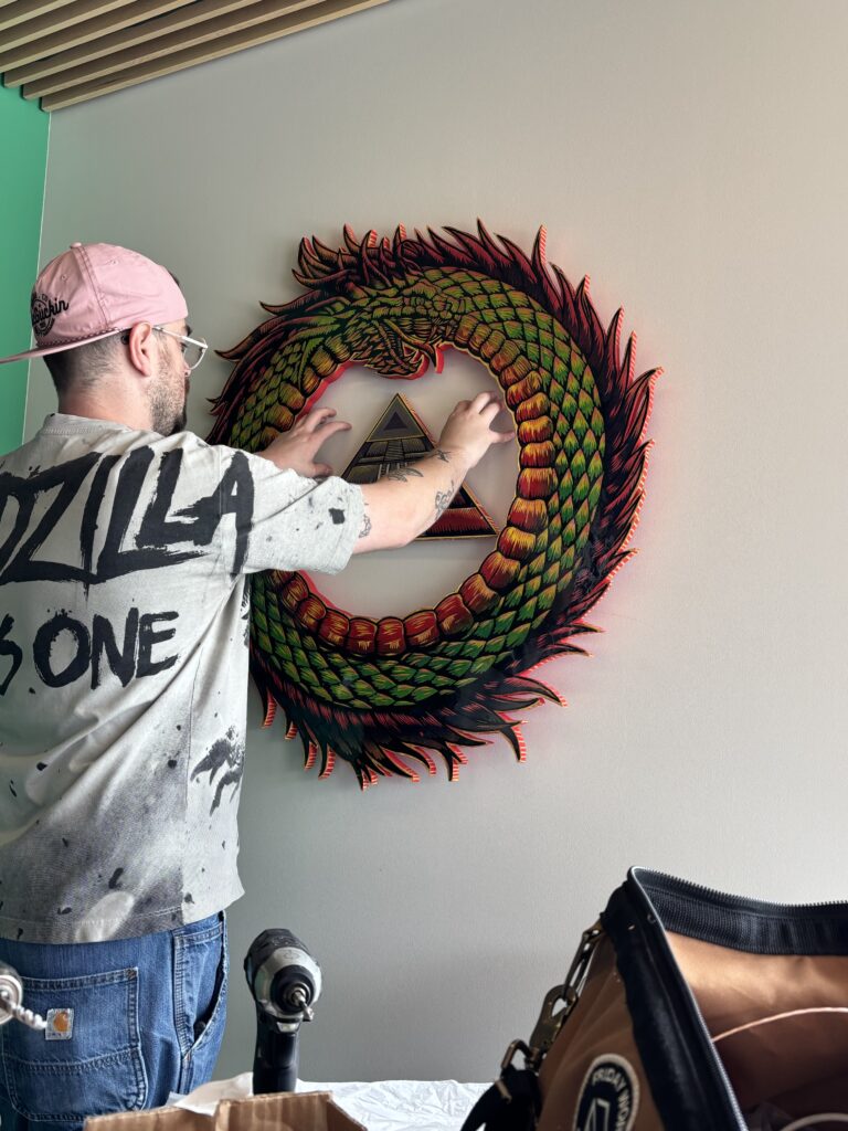 A person installs a large circular wood artwork on a library wall, featuring a serpent-like creature with green and red scales curled around a central pyramid design.