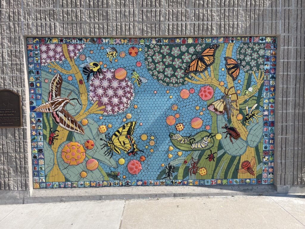 A large mosaic artwork on an exterior wall depicting flora and fauna, including butterflies, moths, bees, a caterpillar and beetles among blue tiled backgrounds and floral clusters, bordered by a colorful strip of small insect and flower tiles.