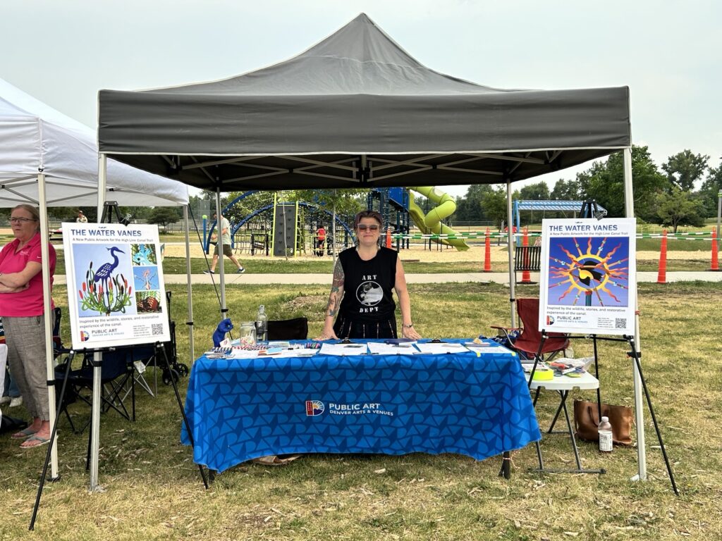 A Public Art staff member stands at an outdoor tent booth displaying posters for "The Water Vanes" artwork, with tables of activity materials and a playground visible behind them.