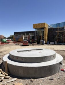 A round concrete sculpture foundation poured at a construction site, with the partially built Westwood Recreation Center and construction equipment visible in the background.