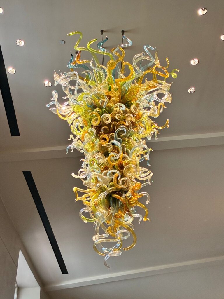 A view looking up at a large blown-glass chandelier of curling amber, green and clear glass forms, glowing under recessed ceiling lights.