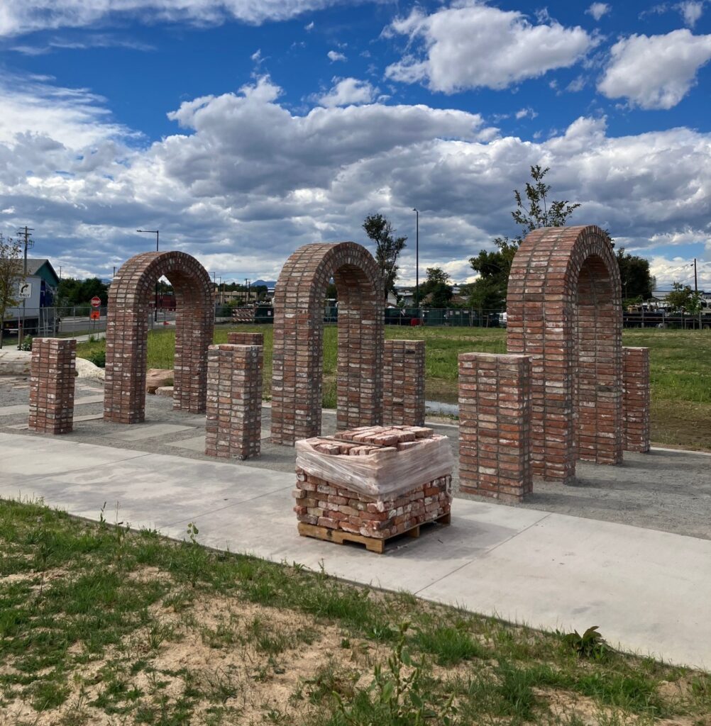 A wider view of three brick arches and pillars lined up along a park path, with a pallet of bricks in front and neighborhood buildings in the distance.